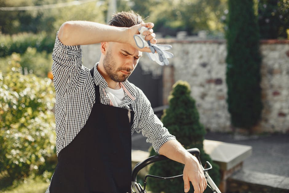 A man and a woman are engaged in a garden maintenance activity within a lush green outdoor space. The man, dressed in a checked shirt, black trousers, and a brown apron, holds a long-handled gardening tool, possibly a rake or a hoes, and appears to be explaining or discussing gardening tasks. The woman, wearing a white t-shirt and distressed jeans with rolled-up cuffs, is holding a small potted plant or flowers, listening attentively. Surrounding them are various garden elements: a pathway running through the garden, a mix of well-maintained grass and slightly overgrown areas with leafy bushes and young trees, and a green mesh fence on the right side. In the background, there is a greenhouse structure with semi-transparent plastic panels, and orange crates stacked against it. The lighting suggests a bright, partly cloudy day, ideal for outdoor gardening activities. This scene illustrates local garden care efforts, showcasing typical tools, plant growth, and the layout of a suburban garden, reflecting the gardening expertise of Gardeners Lambeth.
