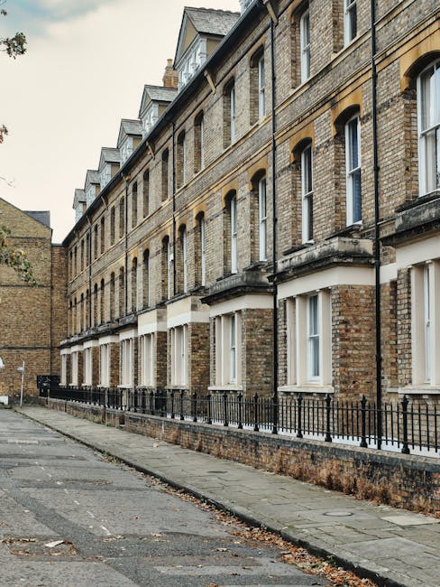 A row of Victorian terraced houses with brick facades and tall, narrow windows, some with white frames, lined along a paved sidewalk. The buildings feature decorative stonework and small balconies with wrought-iron railings under the upper windows. The street is quiet, with a curb separating the pavement from a narrow road that has patches of fallen leaves. In the foreground, a low brick wall runs parallel to the pavement, topped with a simple black metal fence. The environment appears clean and well-maintained, with an overcast sky providing diffuse lighting. Gardeners Lambeth may be involved in garden maintenance or landscaping work related to such traditional terraces, emphasizing care for building exteriors and nearby outdoor spaces in their services, as discussed on the page about Brockwell Park garden maintenance options for Victorian terraces.