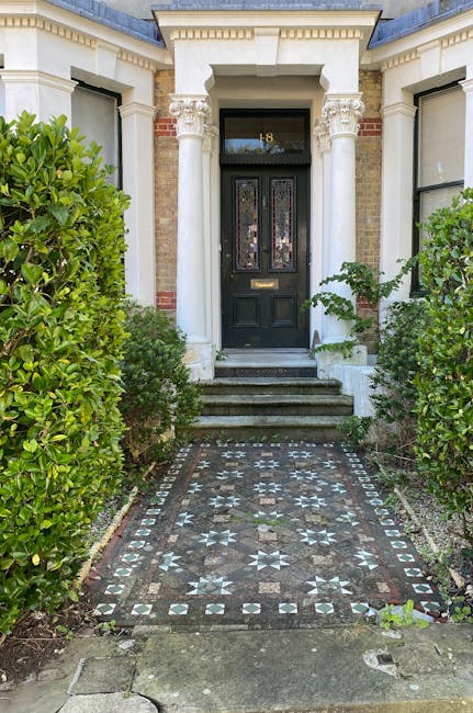 A paved pathway leading to the front door of a Victorian terrace house, flanked by well-maintained green hedges on both sides. The pathway features intricate mosaic tile patterns in earth tones, with a step-up to the black front door, which has decorative stained glass panels and a brass mail slot. Tall white columns support a small portico above the entrance, and the building's facade is composed of red brick with white decorative cornices. Surrounding greenery includes lush bushes, and soft natural light illuminates the scene, highlighting the clean, orderly appearance of the garden area. This setting reflects careful garden maintenance and landscaping work aimed at enhancing the property's curb appeal, as managed by Gardeners Lambeth.