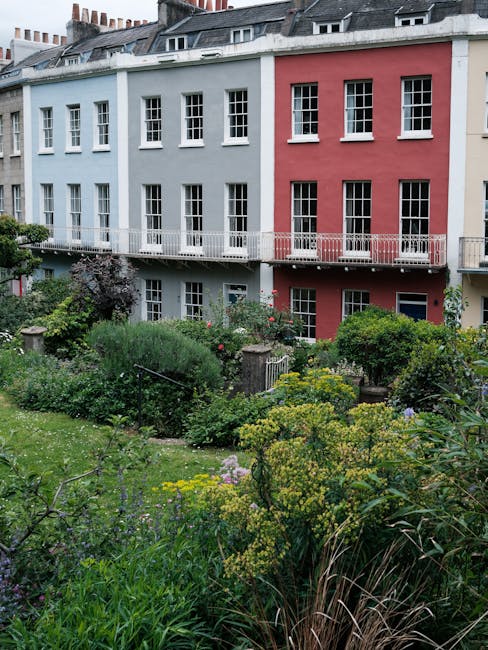A small front garden in an urban residential area, featuring a lush, densely planted garden bed with various shrubs, flowering plants, and ornamental grasses. The garden is surrounded by a low stone wall and has a mix of textures and colors, including green foliage, purple and yellow blossoms, and tall grasses. Behind the garden, there is a row of three-story terraced houses painted in pastel shades of blue and pink, with large white-framed sash windows and small balconies with metal railings. The scene is lit by natural daylight, with a slightly overcast sky, and the overall appearance reflects careful garden maintenance and landscaping work, possibly undertaken by Gardeners Lambeth to enhance small front gardens such as this one.